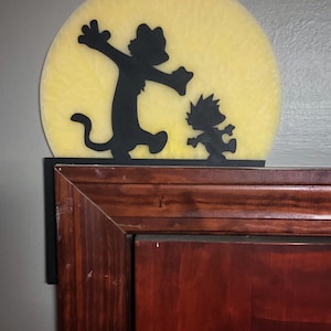 May include: A black silhouette of a cat and a boy running towards each other, set against a yellow, moon-like background. The artwork is mounted on a dark wooden surface, creating a playful and whimsical scene.