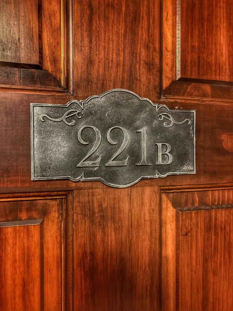 May include: A decorative metal address plaque with the number "221B" in a serif font. The plaque is a dark gray color and is mounted on a wooden door with a rich, reddish-brown finish. The plaque has ornate scrollwork.