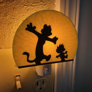 May include: A night light featuring a black silhouette of two cartoon characters on a yellow background. The night light is plugged into a wall outlet and has an on/off switch.