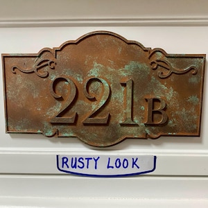 May include: A rectangular address plaque with a rusty brown finish and the numbers "221B" in raised lettering. The plaque has decorative flourishes and a distressed, aged appearance. The text "RUSTY LOOK" is written on a white label below the plaque.