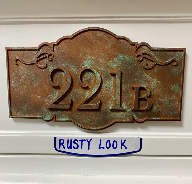 May include: A rectangular address plaque with a rusty brown finish and the numbers "221B" in raised lettering. The plaque has decorative flourishes and a distressed, aged appearance. The text "RUSTY LOOK" is written on a white label below the plaque.