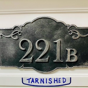 May include: A rectangular, tarnished metal address plaque with the numbers "221B" in silver. The plaque has decorative scrollwork and is mounted on a white surface. The word "Tarnished" is written below the plaque.