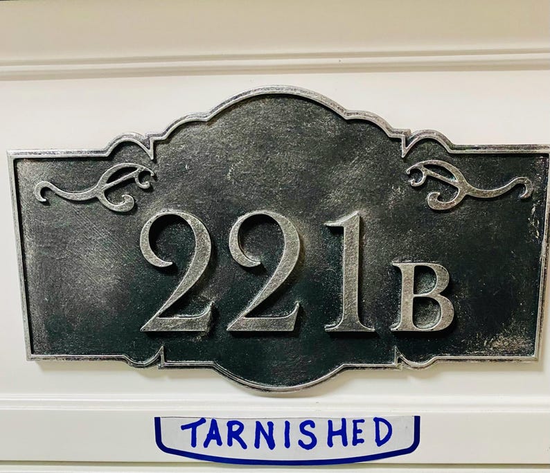 May include: A rectangular, tarnished metal address plaque with the numbers "221B" in silver. The plaque has decorative scrollwork and is mounted on a white surface. The word "Tarnished" is written below the plaque.
