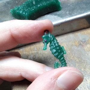 May include: A green wax seahorse figurine with a purple eye. The seahorse is being held by a hand.