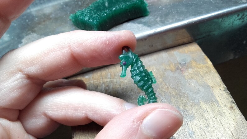 May include: A green wax seahorse figurine with a purple eye. The seahorse is being held by a hand.
