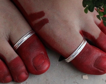 May include: A pair of silver toe rings featuring a simple, polished band design with a subtle textured center line. The rings are worn on the second toes, which are adorned with red pigment, alongside a silver sequined fabric edge.