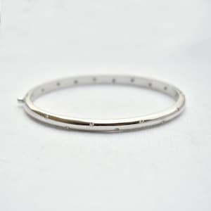 May include: A silver bangle bracelet with small, evenly spaced clear gemstones. The bracelet has a smooth, polished surface and a simple clasp. The jewelry is set against a white background.
