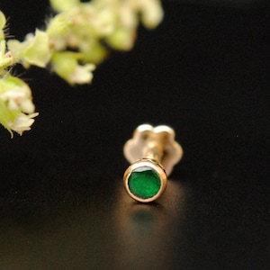 Puede incluir: Un piercing de nariz dorado con una gema verde redonda y brillante. El piercing tiene una parte trasera en forma de flor. La gema tiene aproximadamente 0,25 cm de diámetro, sobre un fondo oscuro.