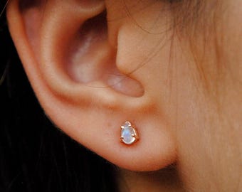Puede incluir: Primer plano de una oreja con un pequeño pendiente de botón en forma de lágrima. El pendiente tiene un engaste de oro y una piedra preciosa azul claro. Es una joya delicada.