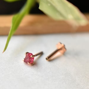 Puede incluir: Primer plano de dos pequeños pendientes de botón de oro rosa. Cada pendiente presenta una gema roja en bruto sujeta por cuatro garras. Los pendientes están colocados sobre un fondo suave y neutro con una hoja verde borrosa.