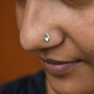 May include: A silver nose stud featuring a pear-shaped, light-colored stone accented with small, clear gemstones. The jewelry is set on a person's nose, with a blurred background.