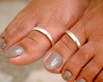 May include: Close-up of two feet with silver toe rings. The toenails are painted a neutral taupe color. The rings are simple bands, reflecting light. The skin tone is medium, and the focus is on the jewelry.