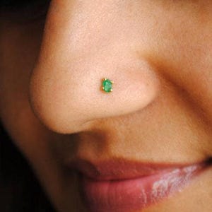 Puede incluir: Primer plano de una nariz con un pequeño piercing de nariz de esmeralda de corte cuadrado engastado en oro. La piedra preciosa verde brillante contrasta con el tono de la piel, resaltando el delicado diseño de la joya. Los labios son visibles.