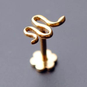 May include: A gold-colored labret stud featuring a stylized snake design. The snake curves upwards, with a post and a flower-shaped base. The jewelry is likely made of gold or a gold-colored metal.