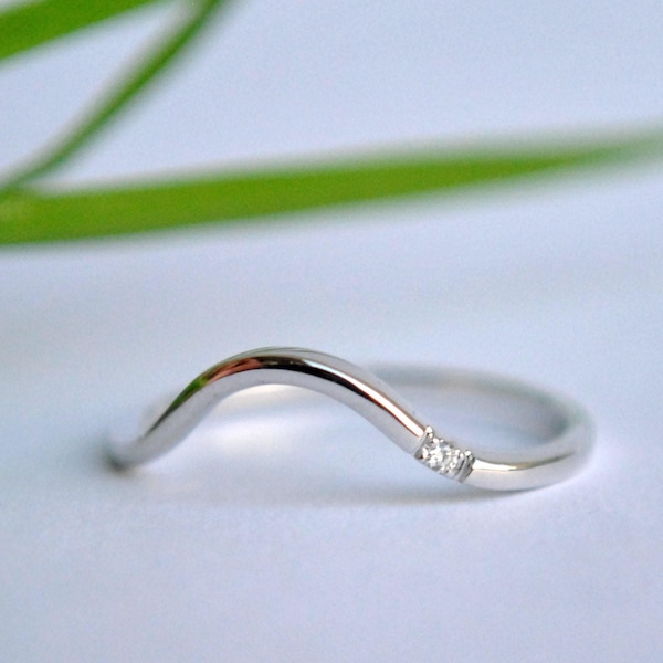 Puede incluir: Un anillo plateado curvado con un diseño delicado. El anillo presenta una forma de onda y está adornado con tres pequeñas gemas brillantes. El anillo se encuentra sobre un fondo suave y borroso, resaltando su elegancia.