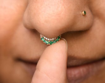 Könnte beinhalten: Ein goldener Septumring mit geschwungenem Design, besetzt mit einer Reihe kleiner grüner Steine und einer zweiten Reihe klarer weißer Steine. Ein kleiner Goldstecker mit einem einzelnen grünen Stein ist am Nasenflügel zu sehen.