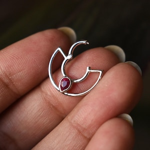 May include: A silver-colored earring with a teardrop-shaped, deep red gemstone. The earring has a unique, open design with curved lines and a hinged clasp. The gemstone is set in the center of the earring, adding a pop of color.