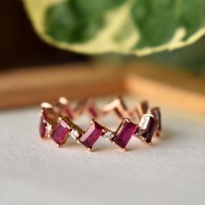 Op de afbeelding: Roségouden ring met afwisselende rechthoekige rode edelstenen en kleine witte diamanten. Het ontwerp van de ring is een herhalend zigzagpatroon, waardoor een doorlopende band van kleur en glans ontstaat. De ring ligt op een wit oppervlak.