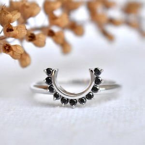 Può includere: Un anello color argento caratterizzato da una fascia curva a forma di ferro di cavallo, decorata con una serie di piccole pietre rotonde nere incastonate lungo l'arco. La fascia metallica è liscia e lucida, in contrasto con le pietre scure.