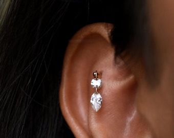 May include: A gold-colored earring with three clear, sparkling stones. The top stone is round, the middle stone is square, and the bottom stone is pear-shaped. The earring is worn in the ear.
