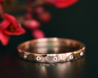 Puede incluir: Un anillo de oro rosa martillado con pequeñas piedras preciosas transparentes. El anillo está enfocado contra un fondo borroso de flores rojas y una superficie oscura. Este anillo es una joya.