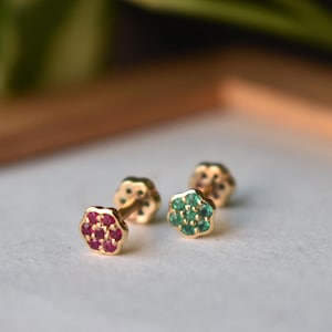May include: Two gold stud earrings featuring a flower-shaped design. One earring is set with seven small round red rubies, while the other earring is set with seven small round green emeralds. Both studs have a thin gold post and a flat back.
