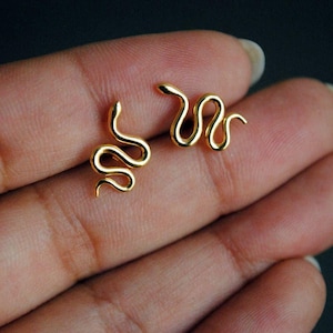 May include: A pair of gold-colored snake-shaped stud earrings. The earrings are small, with a coiled design, and are shown against a neutral background. The earrings are likely made of metal and are a jewelry item.