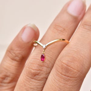 May include: A gold-colored ring with a V-shaped design. A small, clear gemstone is set at the top of the V, and a dangling, marquise-cut, red gemstone hangs below. The ring is on a finger.