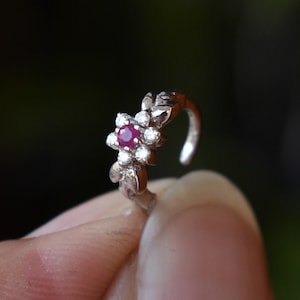 May include: A silver nose ring with a floral design. The central flower has a round, deep red gemstone, encircled by smaller, clear gemstones. The ring is held by a finger against a blurred green backdrop.