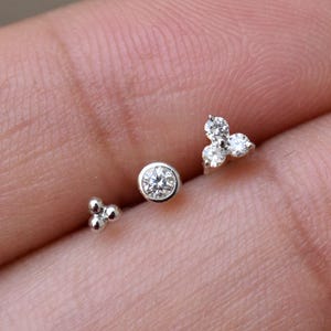 Puede incluir: Tres pendientes pequeños de color plateado. Uno presenta una sola gema redonda y transparente. Otro tiene tres pequeñas gemas transparentes en un grupo. El tercer pendiente tiene tres pequeñas esferas plateadas.