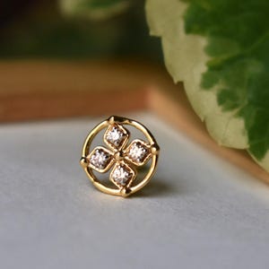 May include: A small, gold-colored stud earring with a four-petal design. Each petal features a sparkling, clear gemstone. The earring is set in a circular frame with a delicate, ornate design. The background is blurred, with hints of green and neutral tones.