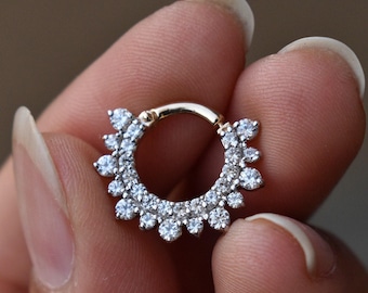 May include: A gold-toned septum ring with a circular design. The ring is adorned with numerous small, clear, round gemstones, creating a sparkling, sunburst effect. The jewelry is held between fingers.