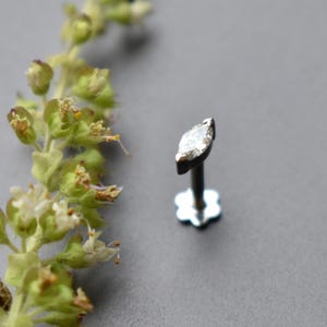 Puede incluir: Un pendiente de botón plateado con una gema transparente con corte marquesa. El poste del pendiente tiene un pequeño respaldo en forma de flor. El fondo es gris liso, con una ramita de flores verdes y amarillas en la esquina superior izquierda.