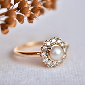 Puede incluir: Un anillo de oro con una perla blanca central rodeada por un halo de pequeños diamantes brillantes. La banda del anillo es de oro pulido. El diseño recuerda a una flor, con la perla en el centro.
