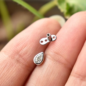 May include: A close-up of two pieces of jewellery. One is a small, silver teardrop-shaped piece with a clear stone and a silver border. The other is a small, silver piece with a post and two holes. The jewellery is on a person's hand.