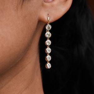 May include: Gold drop earrings with six clear, round gemstones. The earrings hang from a gold hook and are set in a gold and silver bezel. The earrings are displayed against a dark background.