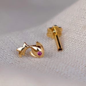 May include: Two gold-colored earrings. One earring is shaped like a fish with a small pink gem. The other earring has a flower-shaped top. Both earrings are on a white surface.