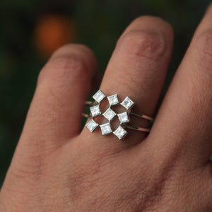 May include: A silver ring featuring a geometric design of square-shaped, clear gemstones. The ring has a double band and the gemstones are arranged in a repeating pattern. The ring is shown on a finger.
