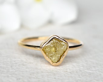 May include: A gold ring featuring a rough-cut, yellow diamond. The diamond is set in a bezel setting, and the band is thin and polished. The ring is displayed on a light-colored surface, with a blurred background.