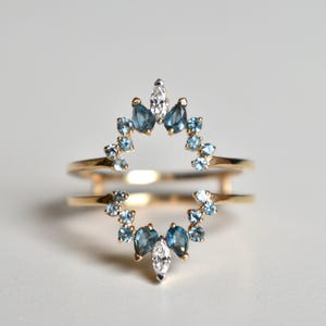 May include: A gold ring with a double band design. The top band features a halo of small, light blue gemstones and larger, darker blue teardrop-shaped stones, with a central white gemstone. Jewelry.
