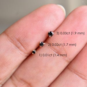 May include: Three small black gemstone stud earrings are displayed. The earrings are set in gold-colored metal. The text on the image indicates the size of each earring: 0.01ct (1.4 mm), 0.02ct (1.7 mm), and 0.03ct (1.9 mm).