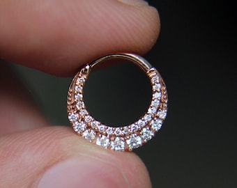 May include: Rose gold septum ring with two rows of clear, sparkling stones. The hinged segment ring is held between fingers against a dark background. The jewelry is designed for nose piercings.