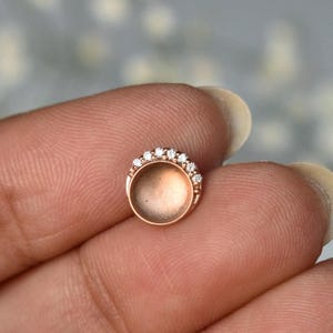 May include: A rose gold-colored stud earring with a circular design. The earring features a row of small, clear gemstones set along the top edge. The stud is held between fingers, showcasing its delicate details and craftsmanship.