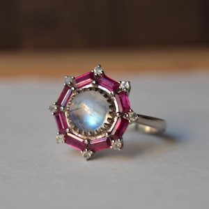 May include: A silver ring with a central, round, iridescent moonstone. It's encircled by a halo of bright pink rectangular gemstones and small, clear accent stones. The ring has a geometric and elegant design.
