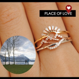 Puede incluir: Tres anillos apilados con diseños de montañas y rayos de sol. El anillo superior es dorado con un diseño de rayos de sol, el anillo central es dorado con un diseño de montaña y el anillo inferior es plateado con un diseño de ola. El texto "PLACE OF LOVE" está en la imagen.