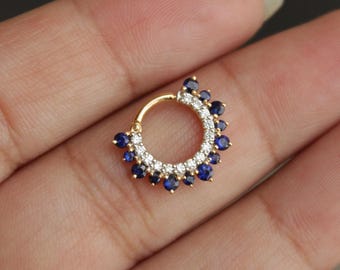 Puede incluir: Un anillo de tabique dorado con una fila de pequeñas piedras preciosas transparentes y una serie de piedras preciosas azul oscuro. El anillo circular está diseñado para piercings en la nariz. Las piedras preciosas están engastadas en un patrón decorativo.