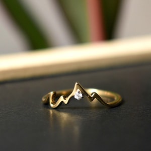 May include: A gold-colored ring with a mountain range design. The ring features a small, clear gemstone set in the center of the mountain peak. The ring is on a dark surface.