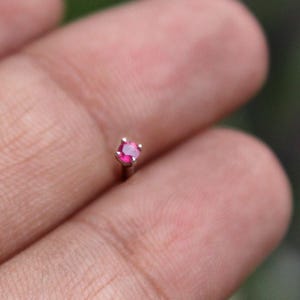 May include: A small, silver nose stud featuring a vibrant, round, ruby-colored gemstone. The gemstone is set with four prongs, and the stud is shown against a blurred background.