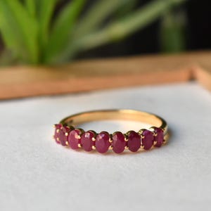 Op de afbeelding: Een gouden ring met een reeks ovaal geslepen, dieprode edelstenen. De edelstenen zijn dicht op elkaar geplaatst, waardoor een doorlopende band van kleur ontstaat. De ring heeft een gepolijste gouden band, die contrasteert met de rode stenen.