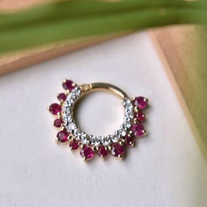 May include: A gold septum ring featuring a circular design. It is embellished with small, round, ruby-red gemstones and clear crystals. The gemstones are arranged around the ring's outer edge and inner circle.